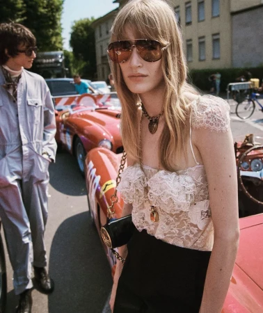 Fashion shot featuring a woman in a lace top and sunglasses beside a red vintage race car, with a man in a jumpsuit in the background.