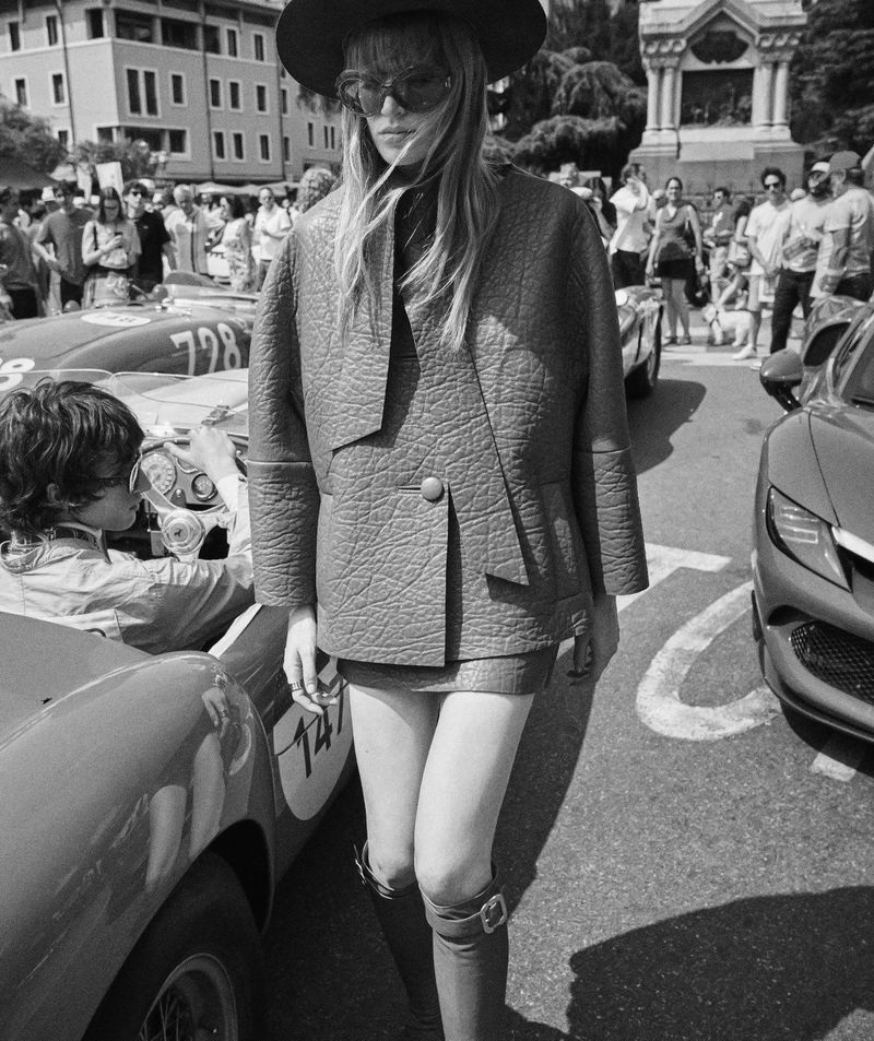 Black and white fashion shot of a blonde woman in a jacket and skirt walking near race cars and a crowd of people on a city street.