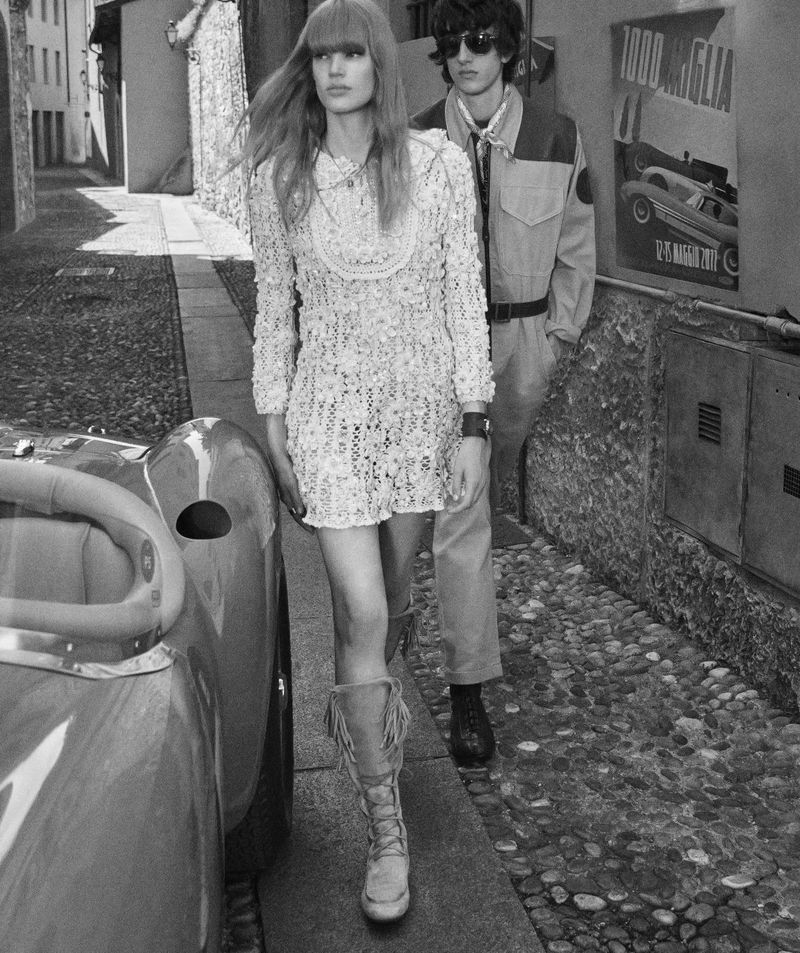 Black and white fashion shot of a woman in a dress and boots walking next to a car, with a man and poster in the background.