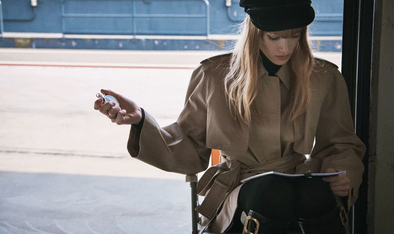 A blonde adult woman in a tan trench coat sitting on a chair and holding a stopwatch in an indoor setting with a neutral background.