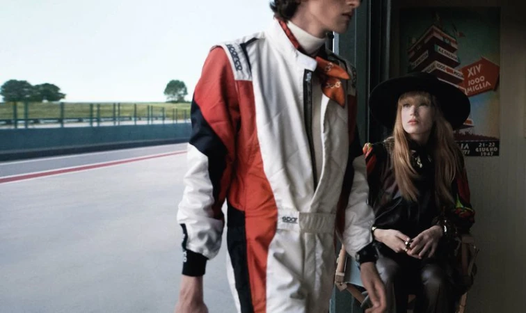 A man in a racing suit stands near a woman in a hat by a poster for the XIV Mille Miglia race, with a race track background.