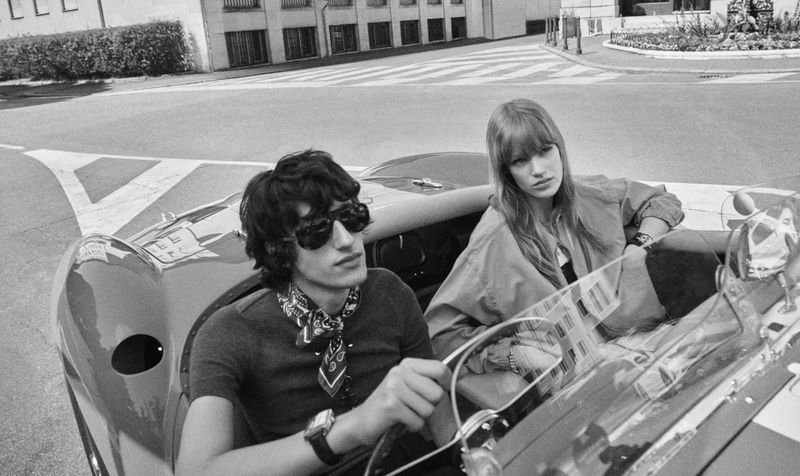 Black and white photo of a stylish couple riding in a classic convertible on a city street; the driver wears sunglasses and a scarf.