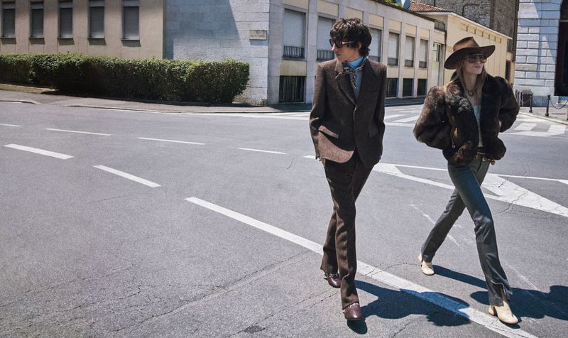 Two fashionably dressed people walk across a crosswalk on a sunny city street, showcasing vintage-inspired outfits with a building backdrop.