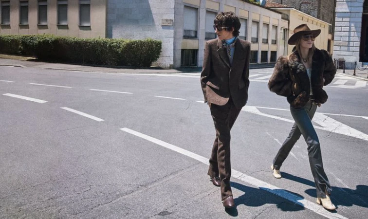 Two fashionably dressed people walk across a crosswalk on a sunny city street, showcasing vintage-inspired outfits with a building backdrop.