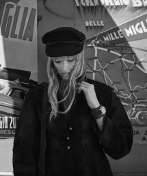Model wearing a dark cap and black jacket standing against a vintage map poster backdrop in a black and white fashion portrait.