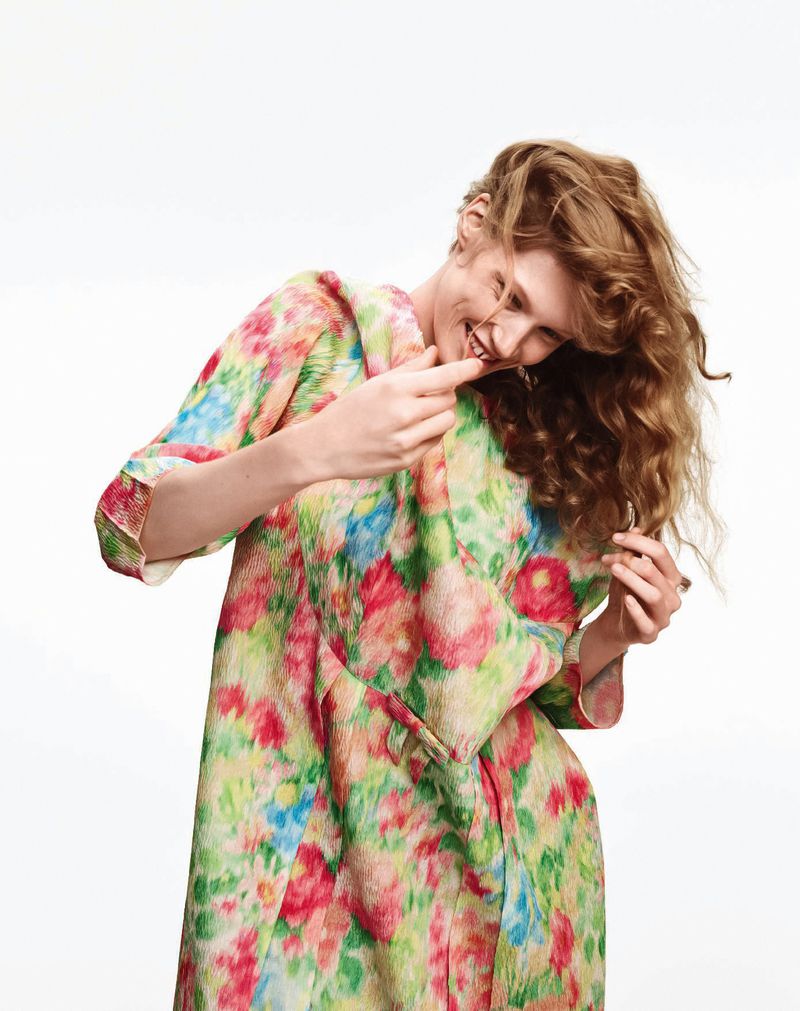 Woman in vibrant floral coat smiles, her hand touches hair, captured against a stark white background in a cheerful fashion image.