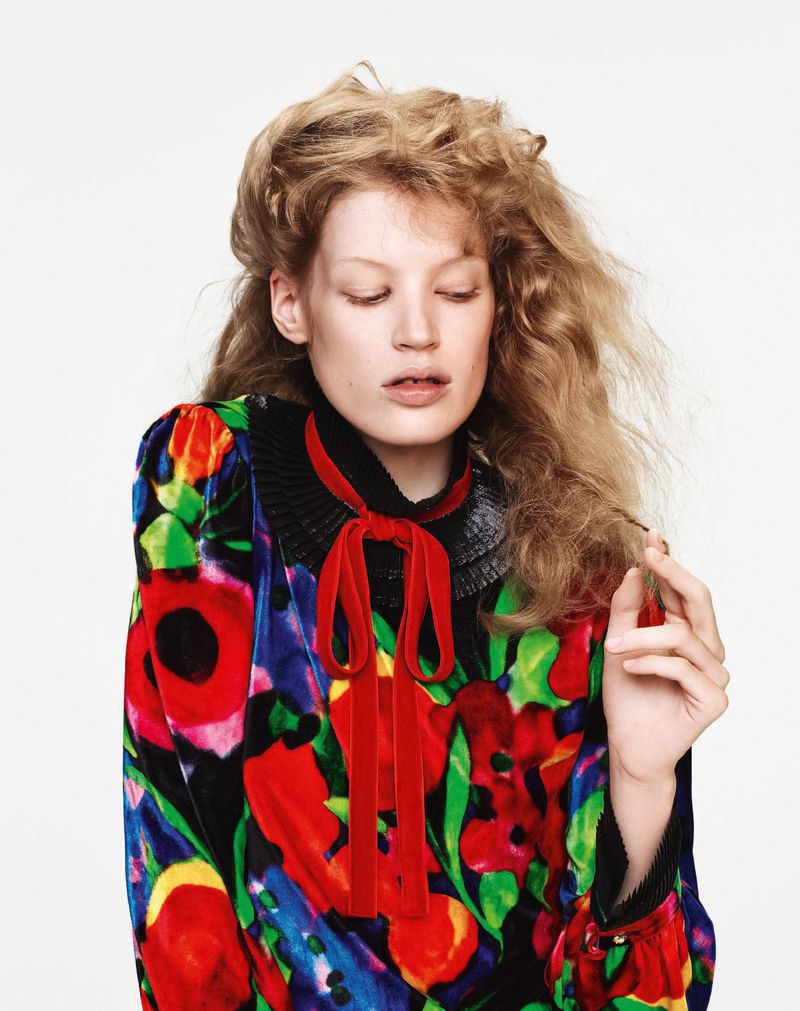 Adult female model with blonde curly hair posing in a flower-patterned, long-sleeve dress and red velvet bow in a studio setting.