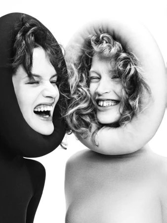 Two people with unique head coverings in a close-up shot. One in a black hood, the other in a white circular frame, both smiling broadly.