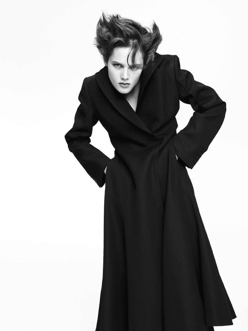Monochrome portrait of a female model wearing a long, dark coat, standing with hands on hips against a bright, neutral backdrop.