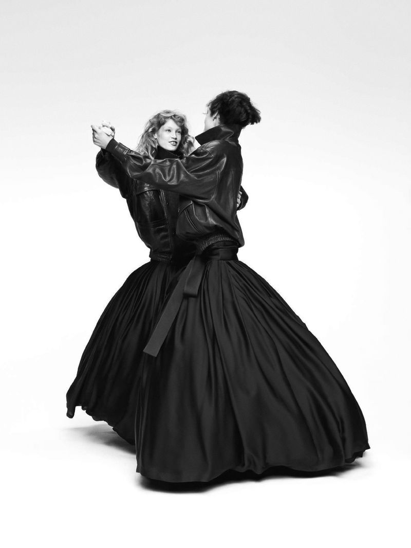 Black and white fashion shot featuring a couple in leather jackets and long skirts, dancing in a bright studio setting. Minimalist aesthetic.