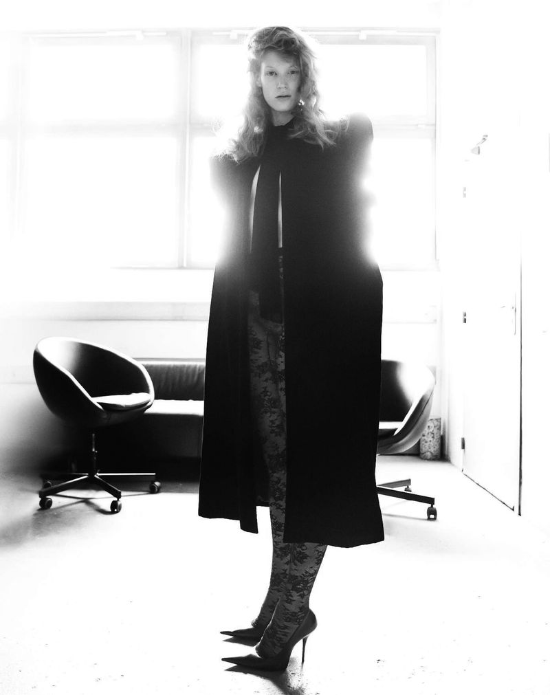 Woman in a long black coat poses in high heels in an indoor setting with a sofa and chair visible in the background.