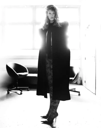 Woman in a long black coat poses in high heels in an indoor setting with a sofa and chair visible in the background.