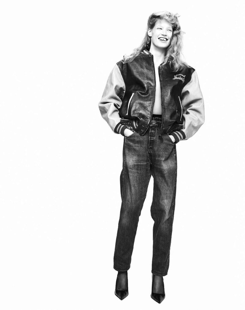 Model laughing in a two-tone leather varsity jacket, dark jeans, and pointed-toe heels against a white studio background.