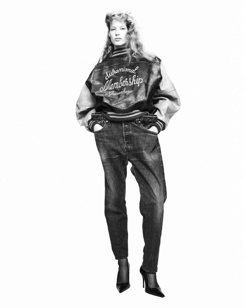 A person in a varsity jacket and jeans poses in a studio shot. The person is wearing black heels and the photo is black and white.
