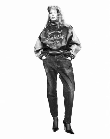 A person in a varsity jacket and jeans poses in a studio shot. The person is wearing black heels and the photo is black and white.
