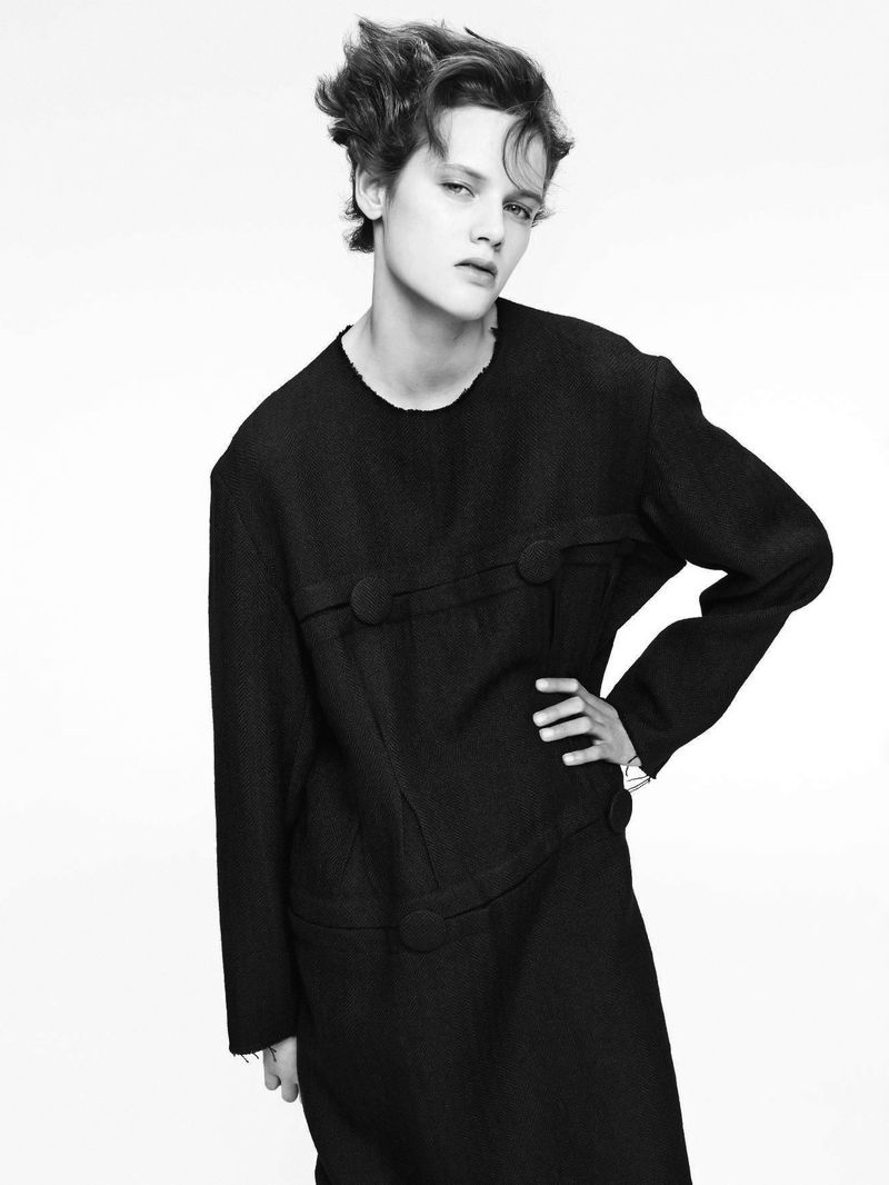 Black-and-white portrait of an androgynous person with tousled dark hair wearing a textured, dark dress, posing against a white background.