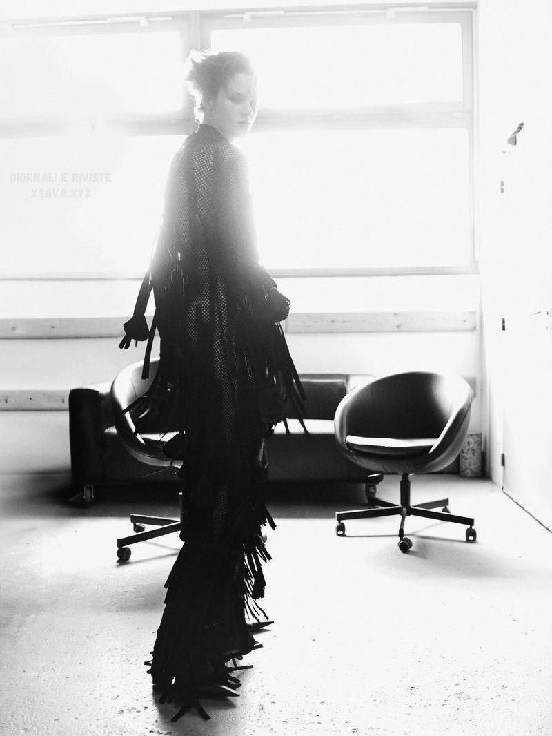 Monochrome studio shot of a woman in a fringed dress, walking past dark furniture and a large window with bright, diffused light.