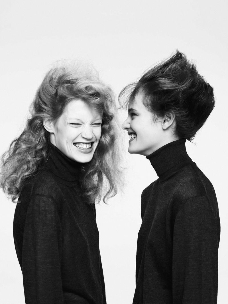 Two cheerful young women with expressive hairstyles, wearing black turtlenecks, share a candid moment against a white backdrop in monochrome.