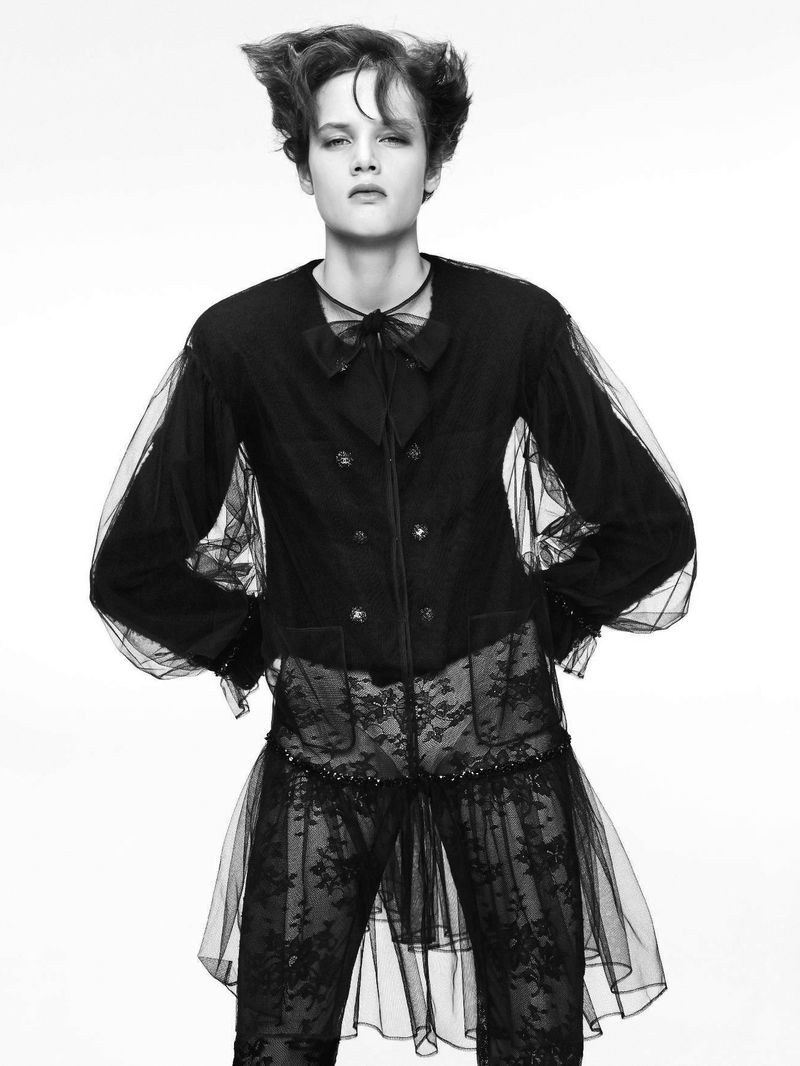 Monochrome portrait of a non-binary person with expressive hair posing in a sheer layered fashion outfit against a neutral background.