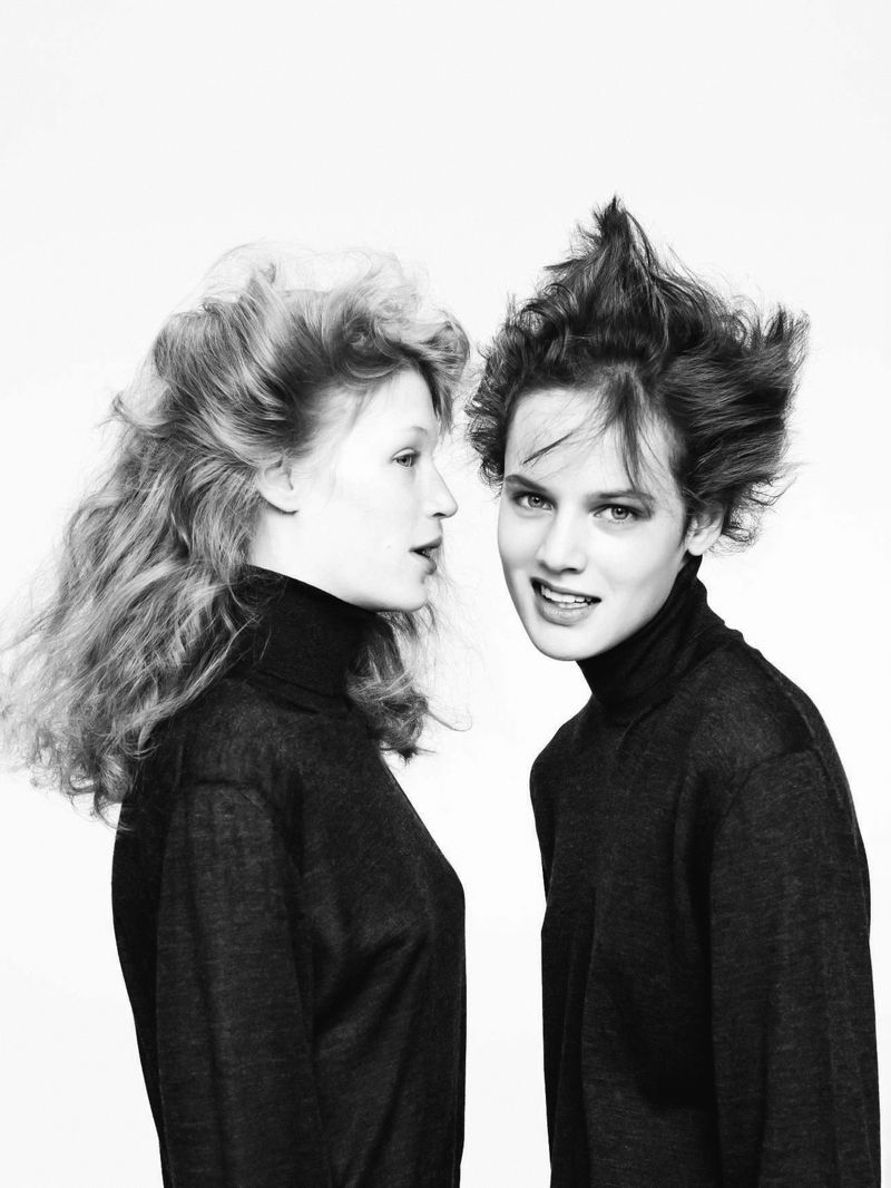 Monochrome portrait of two young adult women with tousled hair wearing black turtlenecks against a bright white background.