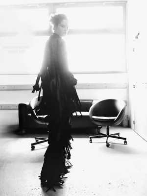 Model wearing a black fringed cape and trousers stands looking over shoulder in a backlit room with office chairs in black and white.