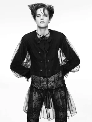 Model with styled short hair posing in a black sheer tulle overlay jacket and lace trousers against a plain white background.