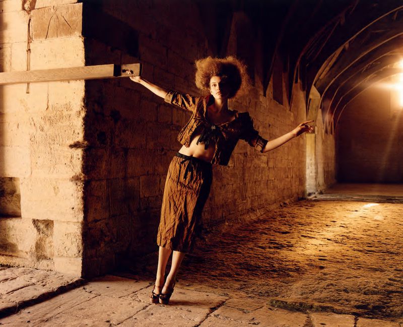 A woman in a brown skirt and jacket leans against a wall in a stone tunnel with light shining at the end, fashion shoot.