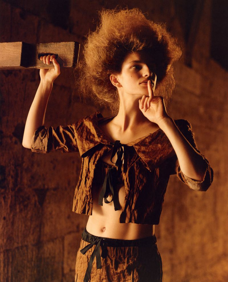 A woman with voluminous hair wears a brown two-piece outfit with black ribbons, posing indoors with a finger to her lips, holding a wooden block.