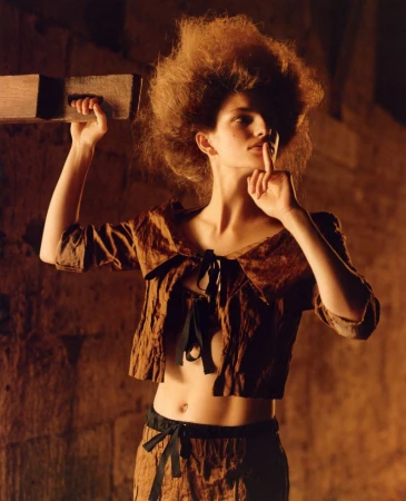 A woman with voluminous hair wears a brown two-piece outfit with black ribbons, posing indoors with a finger to her lips, holding a wooden block.