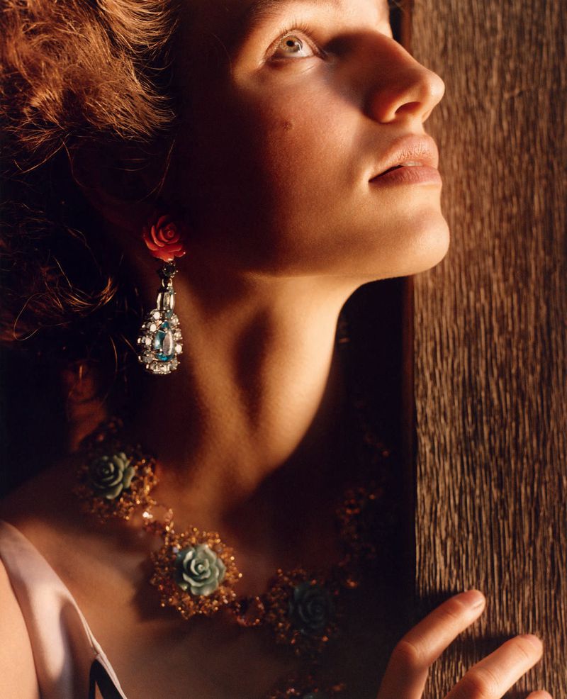 Close-up portrait of a fair-skinned adult female wearing a necklace and earring set with rose and gemstone details, looking up.