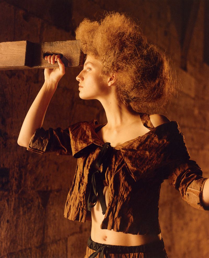 Profile view of an adult woman holding a wooden object, wearing a cropped top with an ornate hairstyle in an indoor environment.