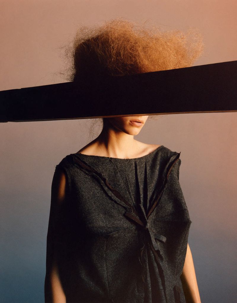 Woman in a grey dress poses in front of a gradient background with a dark horizontal bar obscuring part of her face in a studio shot.
