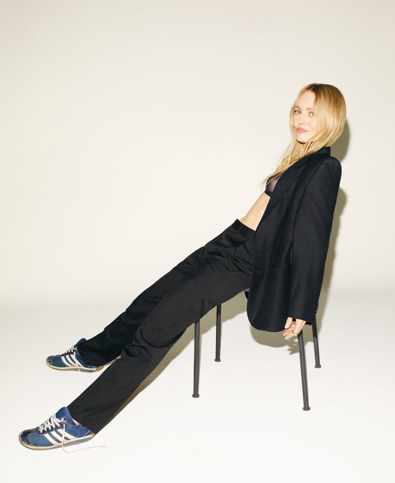 Vanessa Paradis reclining on a black chair wearing an oversized black suit and blue sneakers against a white studio background.