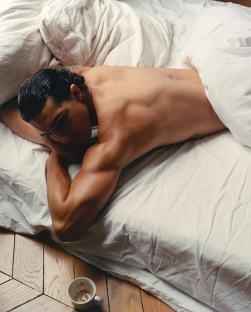Adult male laying in bed under white sheets with empty coffee cup on wood floor in bedroom, top down perspective, indoor setting.