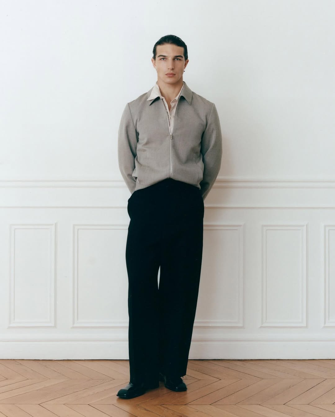 Man in gray jacket and black pants stands in front of white wall with wood floor in full portrait, neutral expression, indoor lighting.