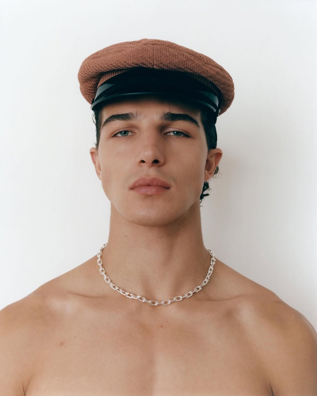 Young man wearing a brown newsboy cap and silver chain necklace against a clean white background