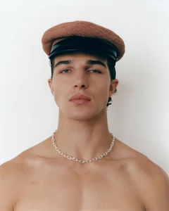 Young man wearing a brown newsboy cap and silver chain necklace against a clean white background