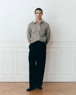 Full-length portrait of a man standing against a white wall in a grey zip jacket and black trousers for Les Hommes Publics.