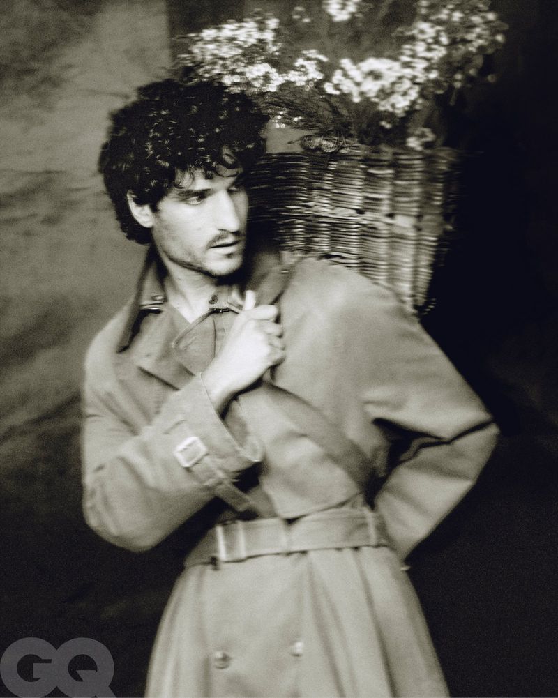 Monochrome portrait of a man in a trench coat carrying a woven basket of white flowers, set against a dark backdrop. Fashion editorial.