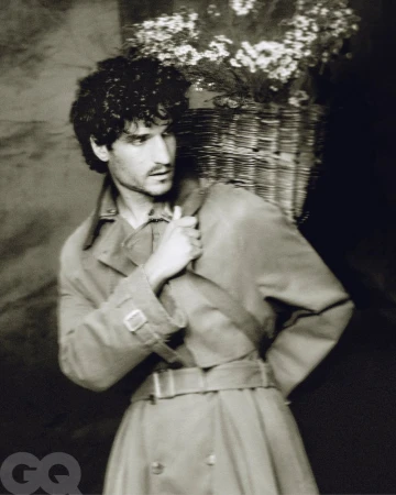 Monochrome portrait of a man in a trench coat carrying a woven basket of white flowers, set against a dark backdrop. Fashion editorial.