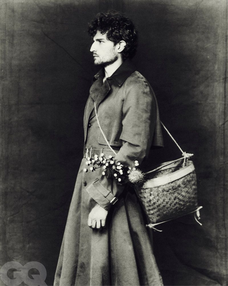 Monochrome portrait of a person in profile wearing a trench coat with a woven bag and flowers, against a dark backdrop.