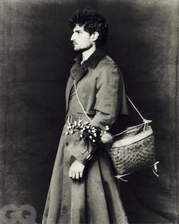 Monochrome portrait of a person in profile wearing a trench coat with a woven bag and flowers, against a dark backdrop.