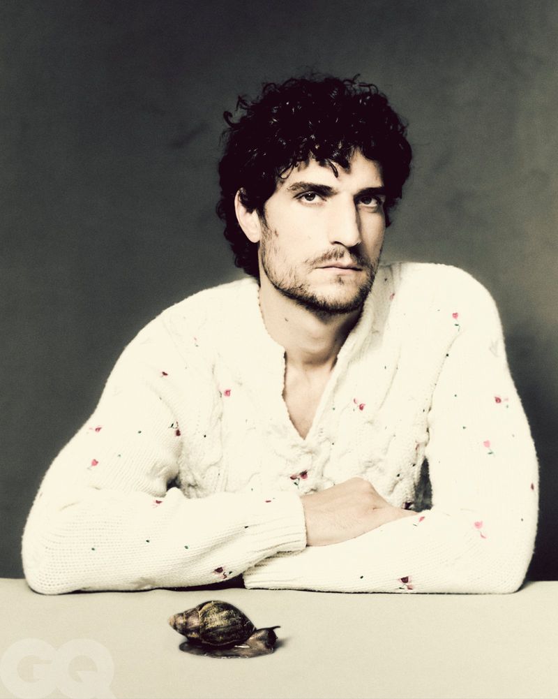 Portrait of an adult male in white patterned sweater posing with a snail on a table against a grey backdrop in soft studio light.