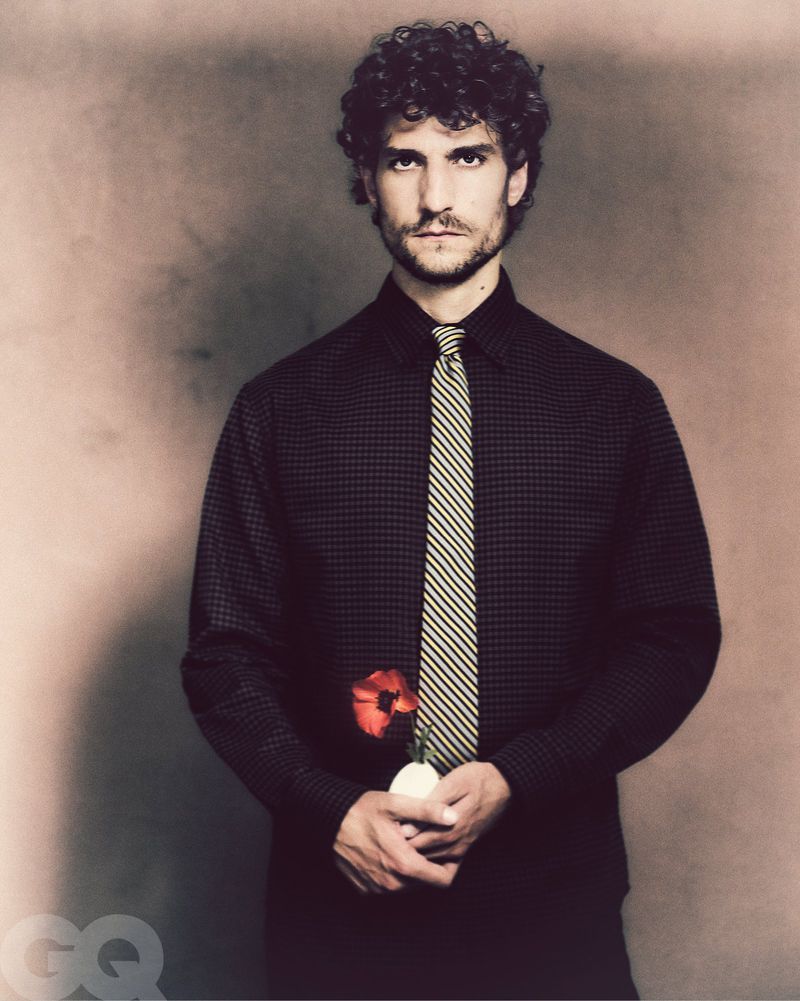 Adult male with curly hair wears a black shirt and yellow tie, holds a red flower against a tan background in a studio portrait.