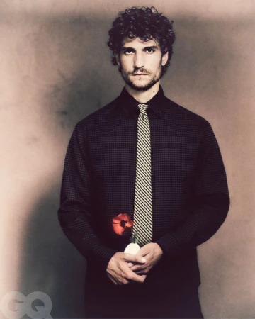 Adult male with curly hair wears a black shirt and yellow tie, holds a red flower against a tan background in a studio portrait.