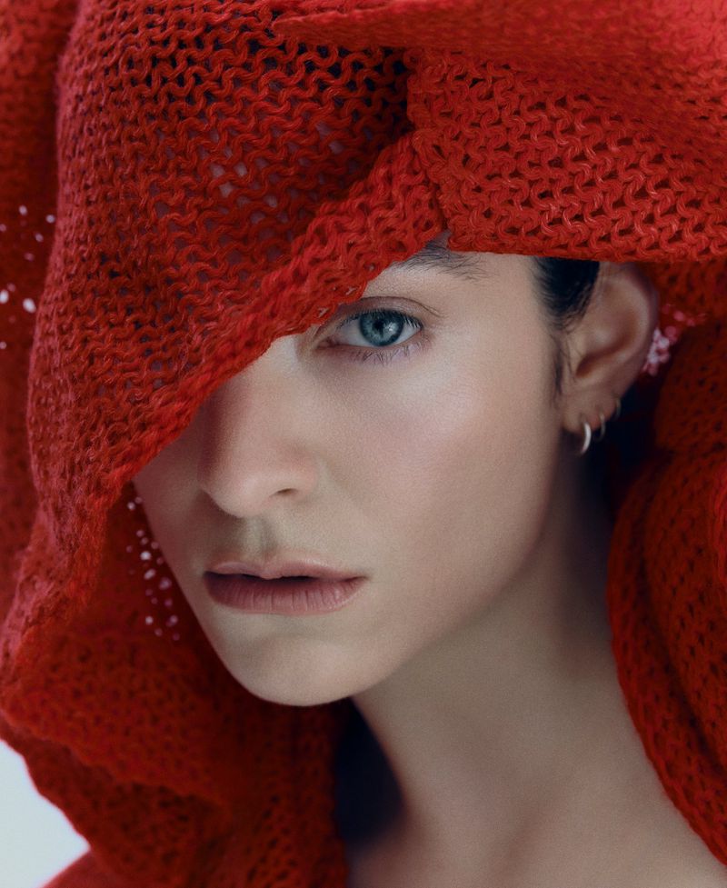 Close-up portrait of singer Lorde draped in a coarse red open-knit hood revealing one eye and silver hoop earrings.