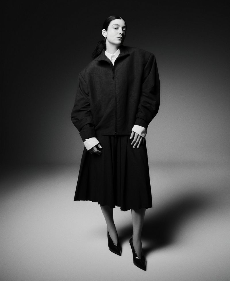 Full-length portrait of a woman in a dark jacket, pleated skirt, and heels against a gray background in a studio fashion shot.