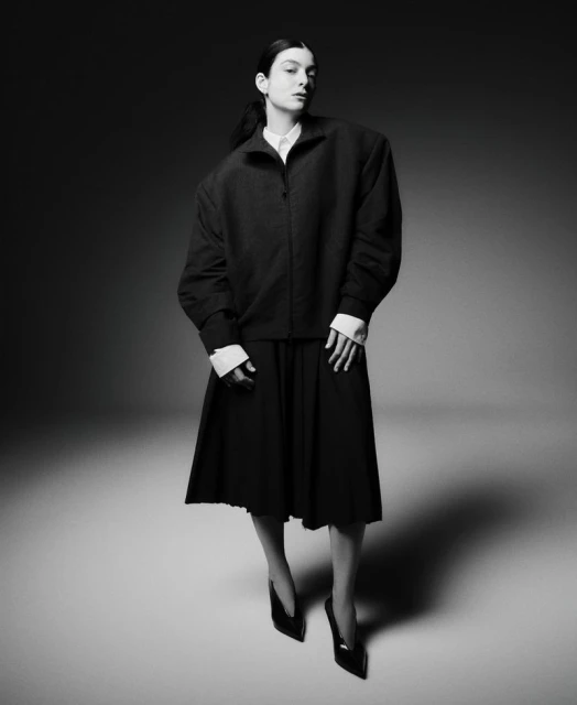 Full-length portrait of a woman in a dark jacket, pleated skirt, and heels against a gray background in a studio fashion shot.