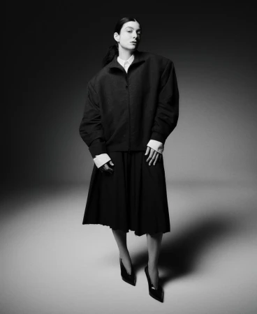 Full-length portrait of a woman in a dark jacket, pleated skirt, and heels against a gray background in a studio fashion shot.
