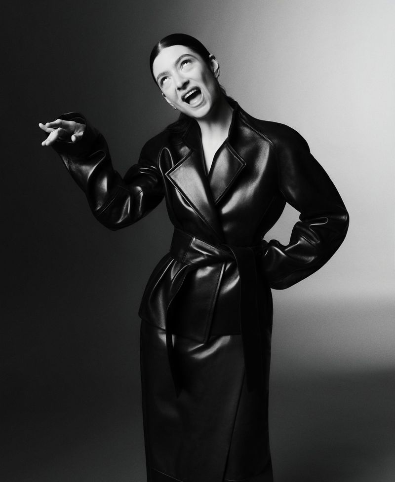 Black and white studio shot of a woman in a glossy black trench coat and matching skirt with her mouth open and arm extended.